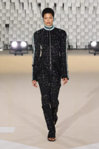 Model walking a fashion show wearing a black sequined zip-front jumpsuit with light blue striped sleeves and open-toed sandals on a neutral runway.