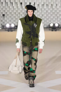 Model walking a runway in an olive-green varsity jacket with white sleeves, patchwork jeans with green star patches, black boots, and a white handbag.