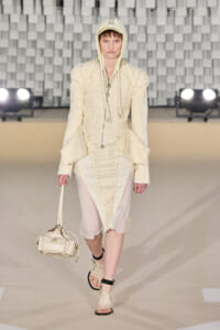 Model walks a cream textured suit with hood on a runway, carrying a small cream handbag and wearing matching sandals.
