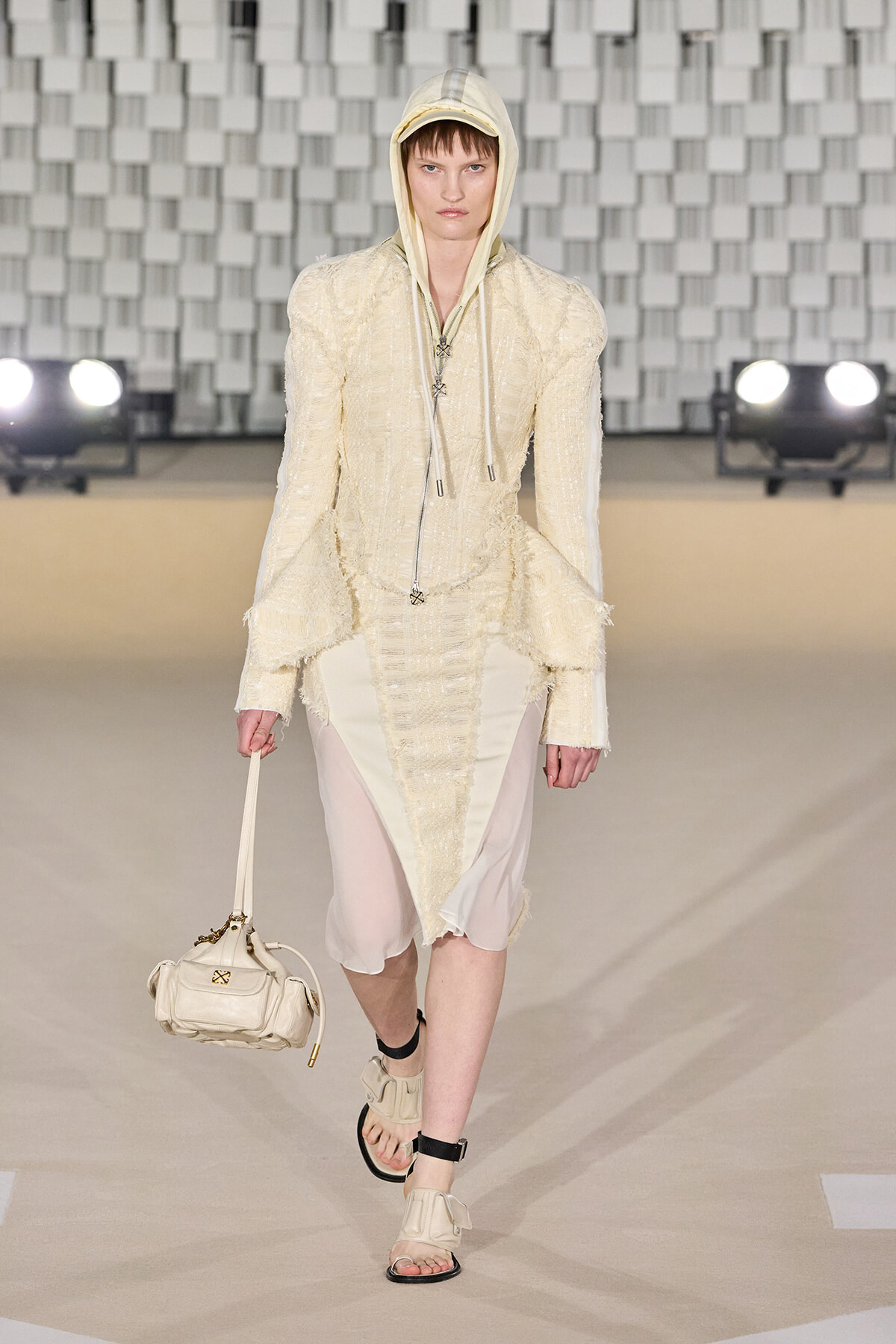 Model walks a cream textured suit with hood on a runway, carrying a small cream handbag and wearing matching sandals.