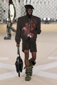 Runway model in a black zip jacket with a red graphic, black shorts, and green chunky strapped boots, carrying a black bag.