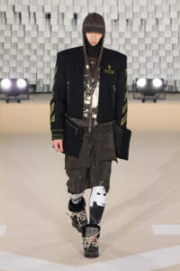 Model walking the runway in an oversized black jacket with neon-yellow patches, layered hoodie, dark graphic shirt, and cargo shorts with patterned leggings and boots.