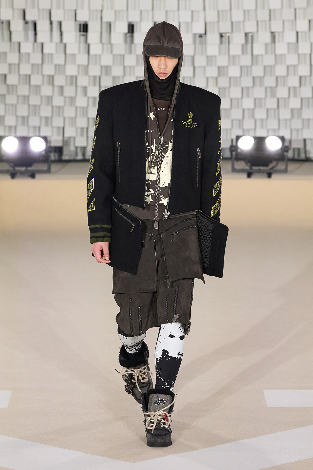 Model walking the runway in an oversized black jacket with neon-yellow patches, layered hoodie, dark graphic shirt, and cargo shorts with patterned leggings and boots.