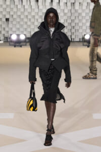 Model on a runway wearing an oversized black hooded jacket with a sequined black skirt and a yellow-and-black striped handbag.