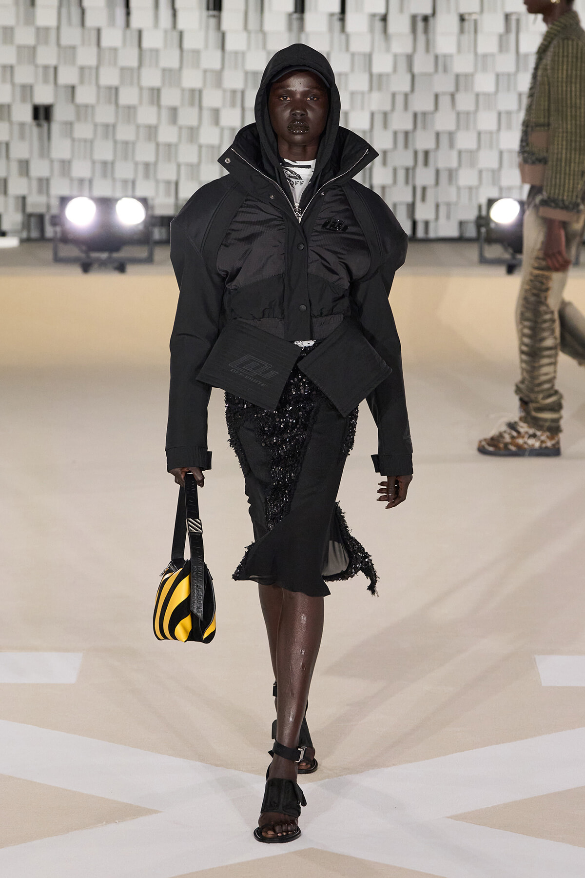 Model on a runway wearing an oversized black hooded jacket with a sequined black skirt and a yellow-and-black striped handbag.