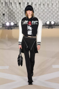 Model walking a runway in a black-and-white varsity jacket with a hooded black cap, graphic tee, black pants, and a black handheld bag.