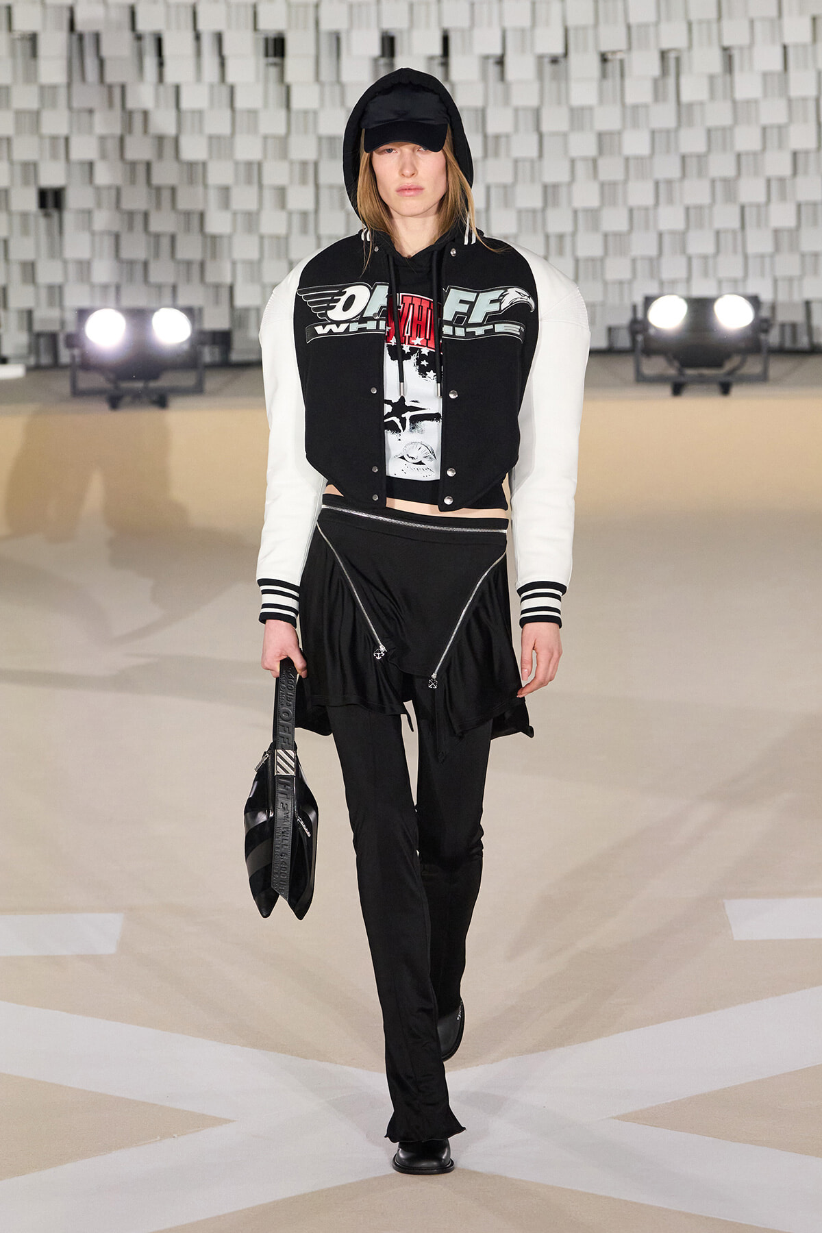 Model walking a runway in a black-and-white varsity jacket with a hooded black cap, graphic tee, black pants, and a black handheld bag.