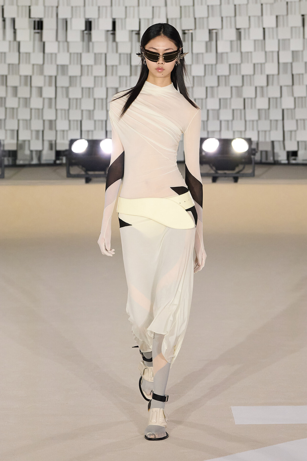 Model walking a runway in a cream sculptural dress with sheer sleeves and futuristic belt, wearing angular sunglasses.