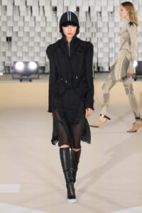 Model walking a fashion runway in an all-black textured outfit: hooded zip jacket, sheer skirt, and knee-high boots.