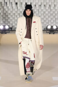 Runway look: a male model in a cream long coat with star patches, black hood, and burgundy knit top with graphic white shorts, rugged boots.
