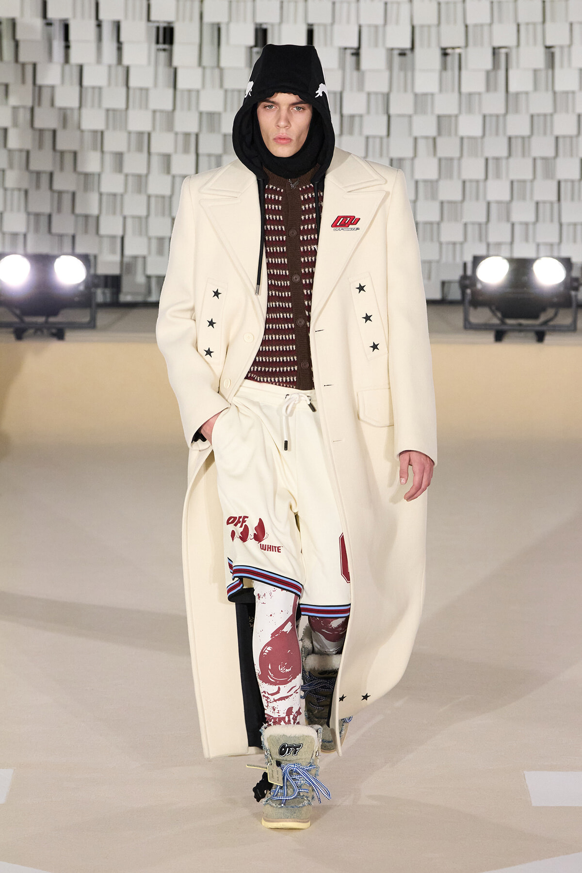 Runway look: a male model in a cream long coat with star patches, black hood, and burgundy knit top with graphic white shorts, rugged boots.