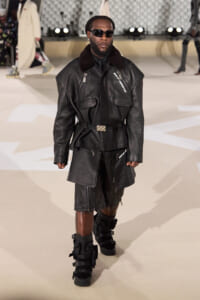 Male model on a runway wearing an oversized black leather jacket with zippers, a matching belt, and knee-length leather shorts with strap boots, plus dark sunglasses.