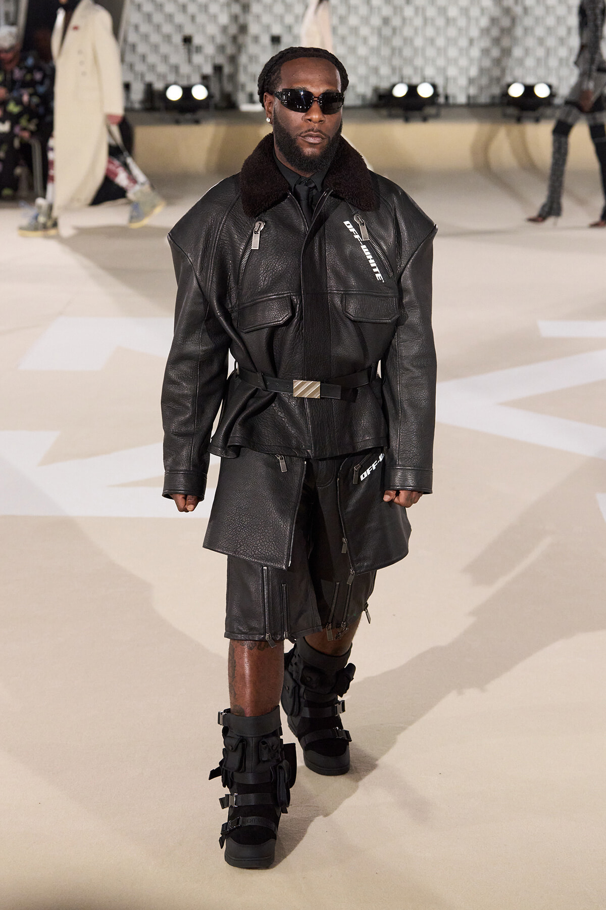 Male model on a runway wearing an oversized black leather jacket with zippers, a matching belt, and knee-length leather shorts with strap boots, plus dark sunglasses.