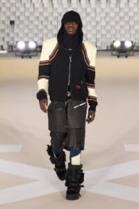 Male model walks a runway in futuristic streetwear: black hoodie under a cream-shouldered jacket with striped sleeves and oversized black shorts with zippers and straps, plus leg braces on both legs.