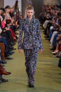 Model walking the runway in a matching floral suit with a double-breasted blazer; audience seated along the sides watching.