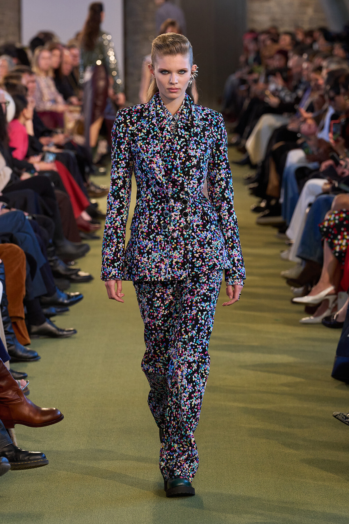 Model walking the runway in a matching floral suit with a double-breasted blazer; audience seated along the sides watching.