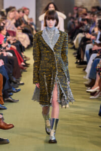 Model walking a runway in a textured dark yellow and black coat with a silver fringe-trimmed scarf and matching fringe skirt details; metallic boots.