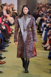 Model walking a fashion show in a burgundy tweed coat with silver-detailed lapels, gray sash, belt, black boots, and a red handbag.