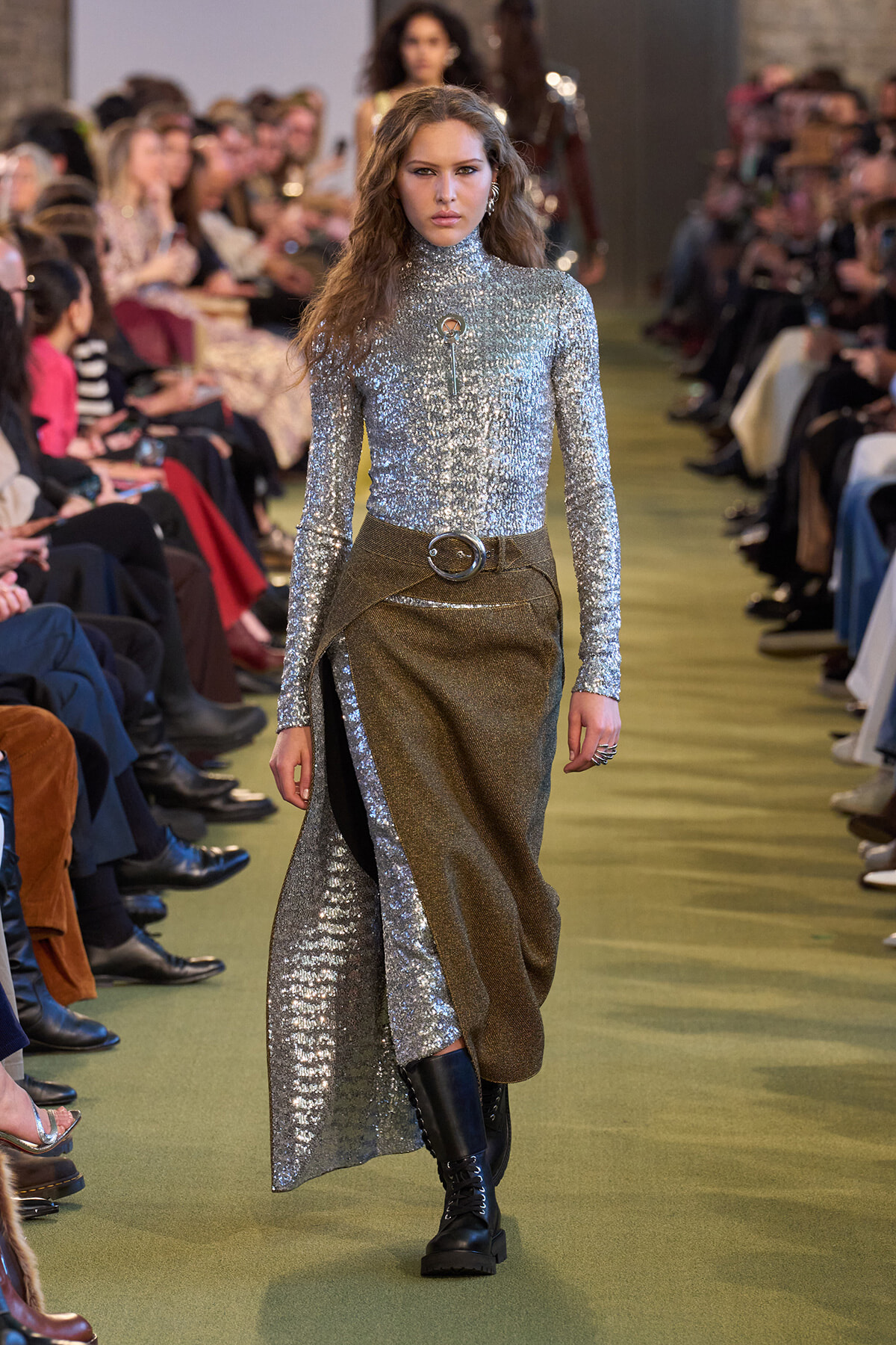 Model walking a runway in a silver sequin turtleneck and metallic wrap skirt with a wide belt and black boots; audience blurred in the background