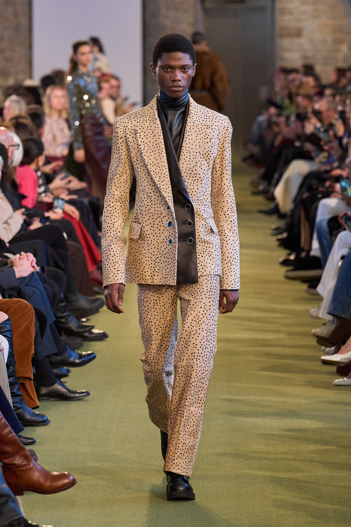 Model on a runway wearing a beige polka-dot suit with a black leather inner layer while the audience watches from the sides.