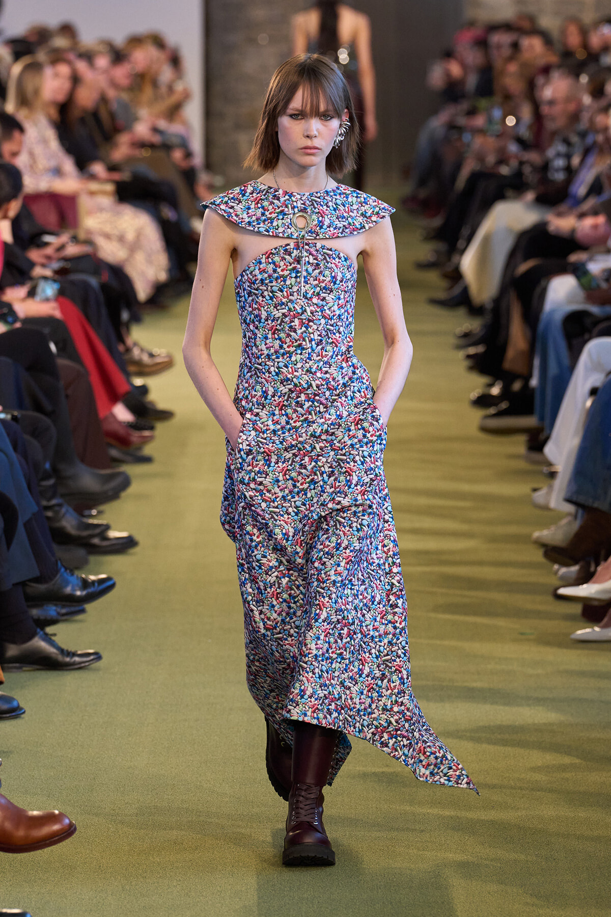 Runway model in a sleeveless, multicolored speckled dress with a collar and cutout detail, paired with burgundy boots.