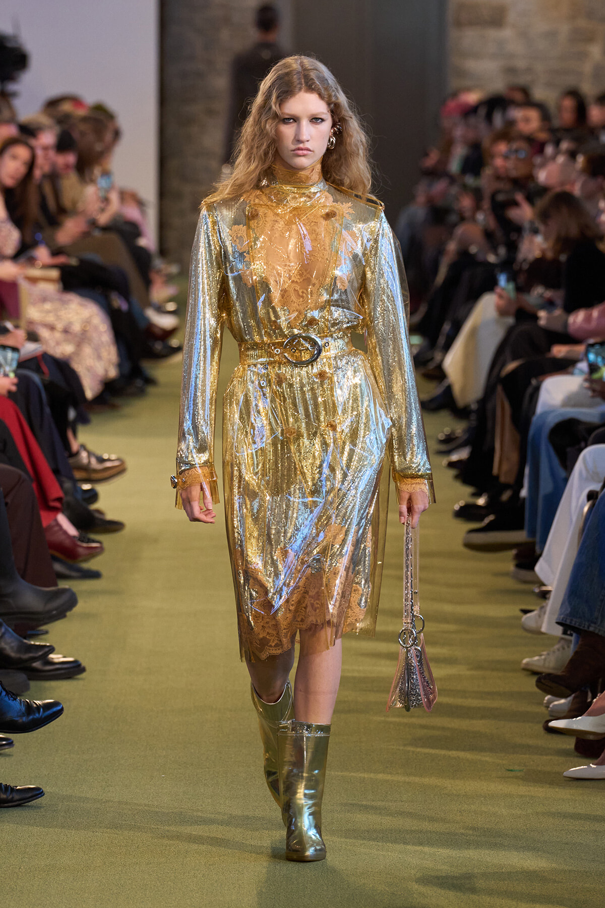 Model walks runway in a metallic gold, translucent trench dress with lace underlayer and matching belt, knee-high boots, carrying a pink metallic bag amid an seated audience.