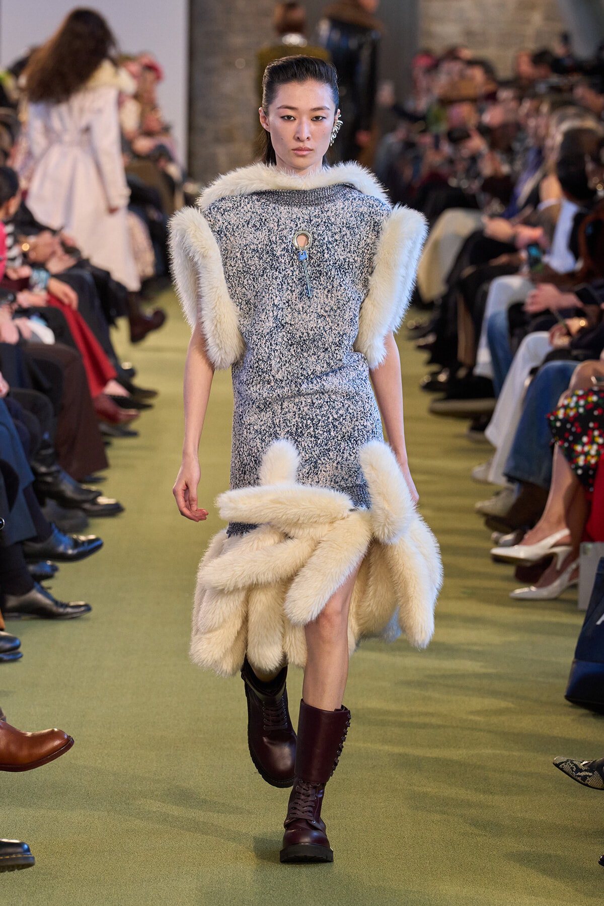 Model walks a runway wearing a textured blue dress with large cream fur trim on the shoulders and hem, paired with burgundy lace-up boots.