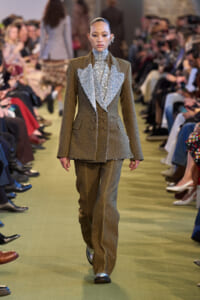 Female model walks a runway in a textured bronze-brown suit with a large silver, ruffled bib-front; audience seated along the catwalk.