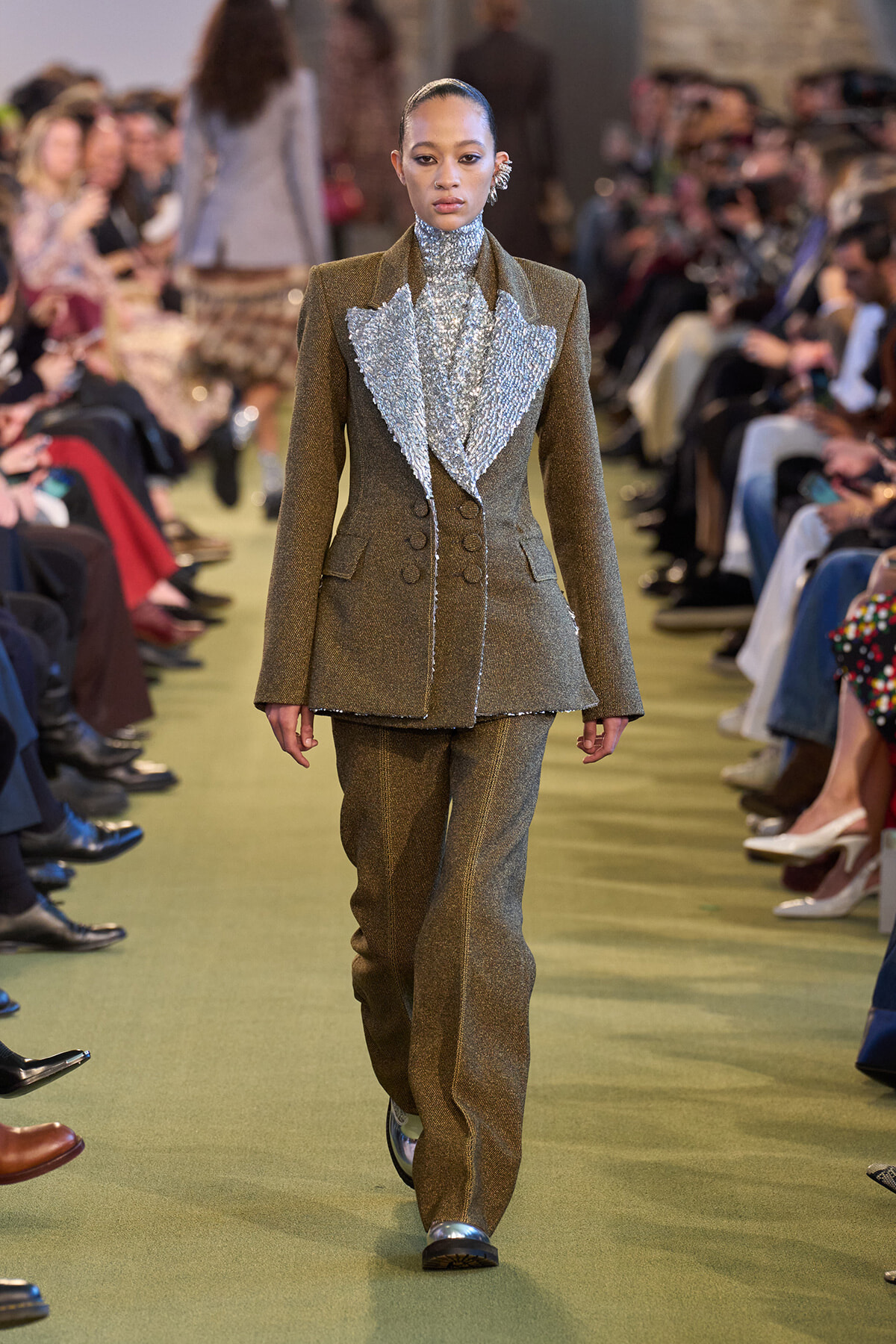 Female model walks a runway in a textured bronze-brown suit with a large silver, ruffled bib-front; audience seated along the catwalk.