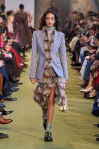 Model walks a fashion runway in a light blue blazer with a fur-trimmed skirt and metallic turtleneck. She wears sparkly socks and black shoes.