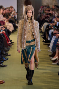 Model walks a runway in a beige polka-dot knit coat with metallic tinsel hood, teal fur-trimmed skirt, and black combat boots.