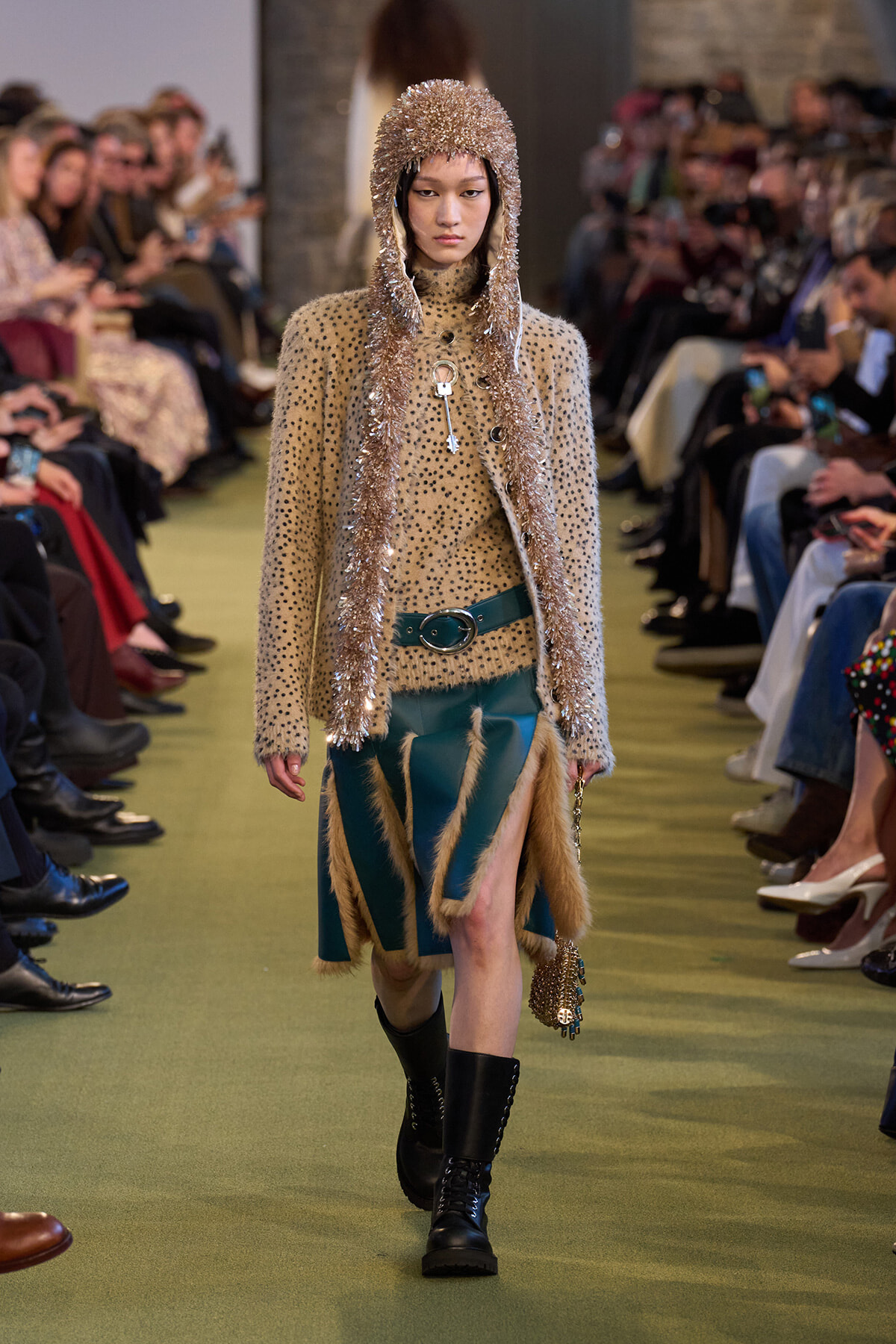 Model walks a runway in a beige polka-dot knit coat with metallic tinsel hood, teal fur-trimmed skirt, and black combat boots.