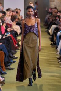 Model walking a runway in a purple sequin top and gold metallic skirt, surrounded by seated audience.