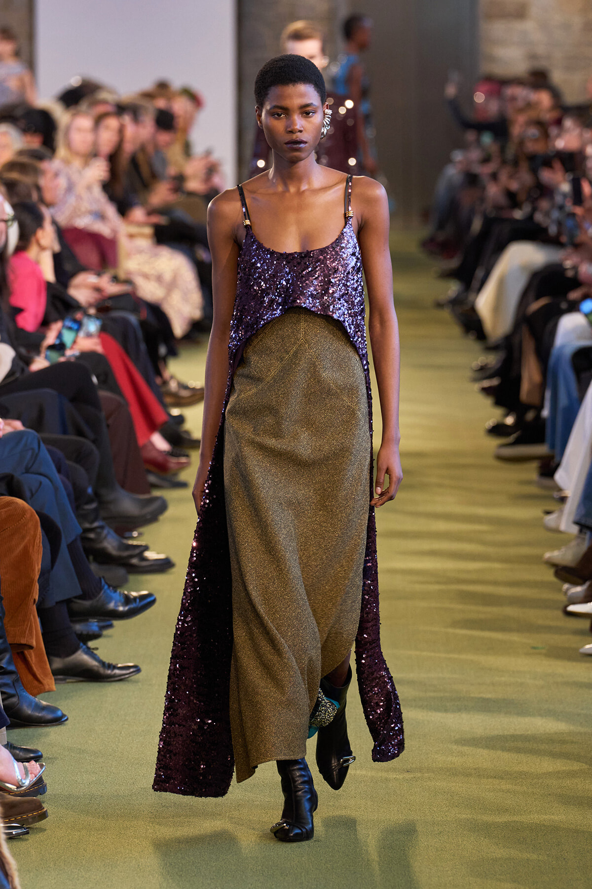 Model walking a runway in a purple sequin top and gold metallic skirt, surrounded by seated audience.