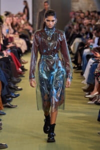 Model walking the runway in a translucent metallic trench coat over a dark outfit, surrounded by seated audience in focus on sides.