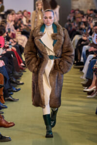 Model walks the runway in a long brown fur coat over a cream outfit with teal trim and a green belt; audience watches in the background.