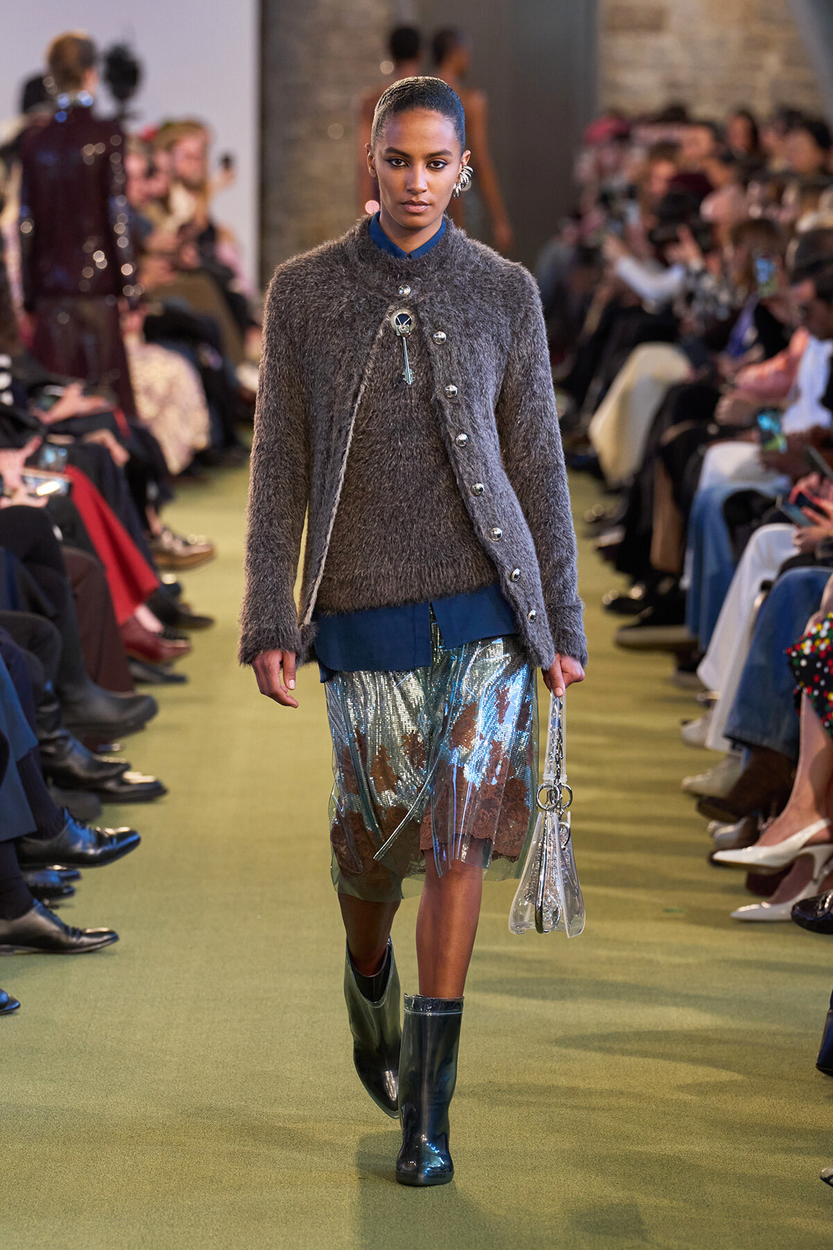 Model walks a fashion runway in a fuzzy gray cardigan over a blue blouse and metallic fringe skirt, with blue boots and a clear bag in hand.