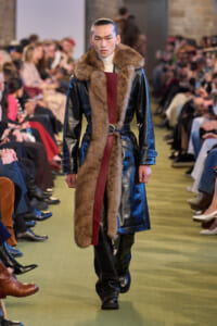 Male model walks runway in a long black leather coat with brown fur trim and a burgundy front panel, audience on both sides.