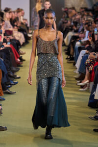 Model walking a runway in a sparkling two-piece outfit: a glittery top with thin straps and a layered sequin skirt over teal glitter pants, audience on both sides
