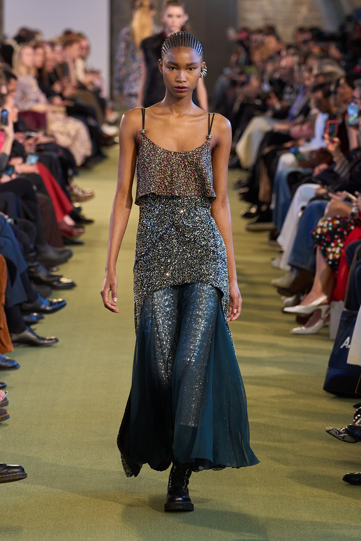 Model walking a runway in a sparkling two-piece outfit: a glittery top with thin straps and a layered sequin skirt over teal glitter pants, audience on both sides