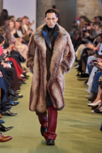 Model walking a runway in a long multicolored fur coat with burgundy leather pants and black boots, audience in the background.