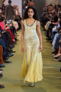 Model walks a runway in a pale yellow sequined gown with a sheer tulle skirt and metallic shoes.