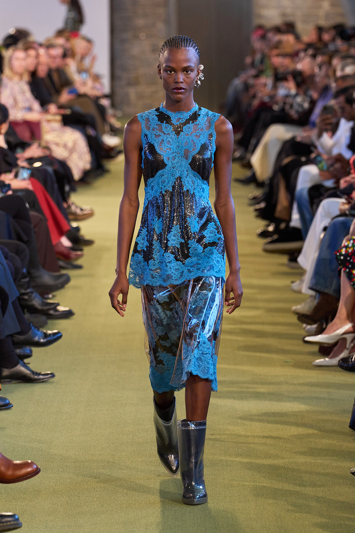 Model walks a runway in a cobalt blue lace dress with sheer panels and knee-high blue boots, audience on both sides watching.