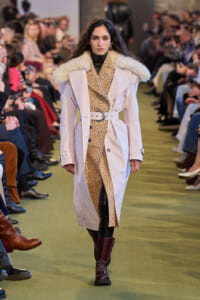 Model walks a fashion runway in a pale coat with a plush fur collar over a beige polka-dot outfit, cinched with a wide white belt and burgundy boots.
