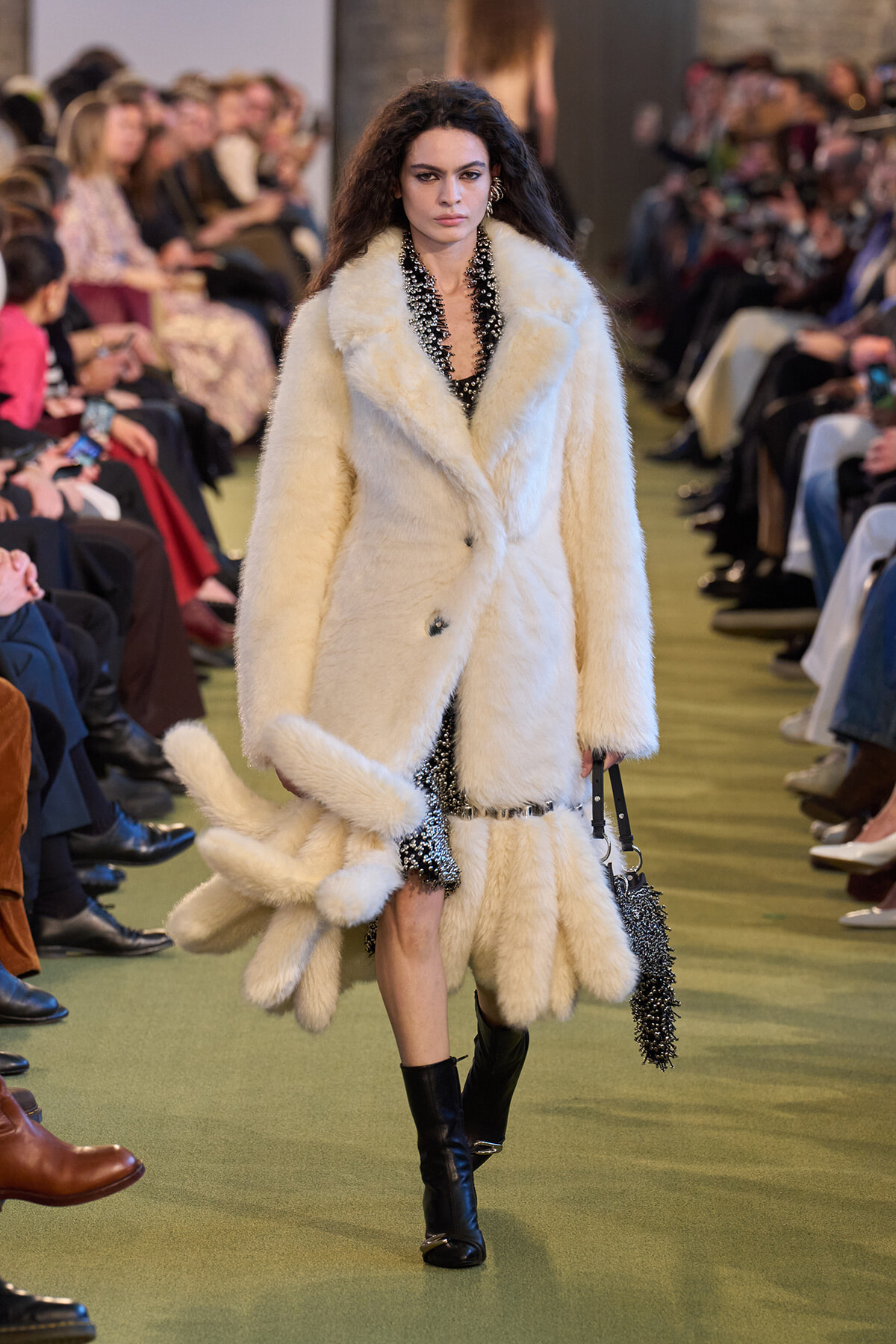 Model walks the runway in a white fur coat over a black sequined dress, knee-high black boots, and a black beaded purse. Audience lines the catwalk.