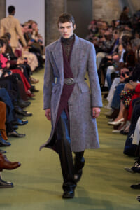 Model walks down runway wearing a long gray textured coat belted at the waist over a burgundy inner layer and black leather pants; audience watches.