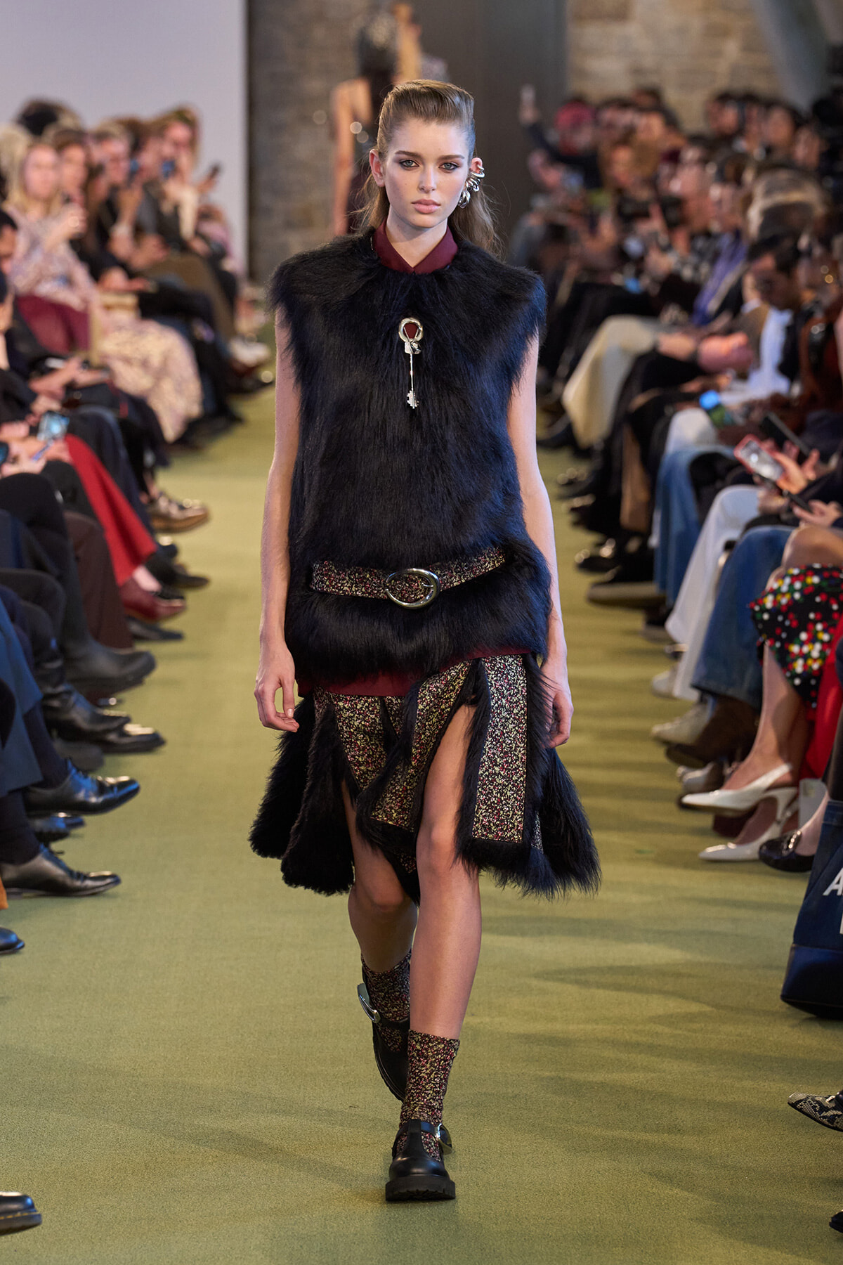 Model walking a fashion runway in a black faux-fur sleeveless top with a metallic belt and glittery panels beneath a fringe skirt; audience blurred in the background.