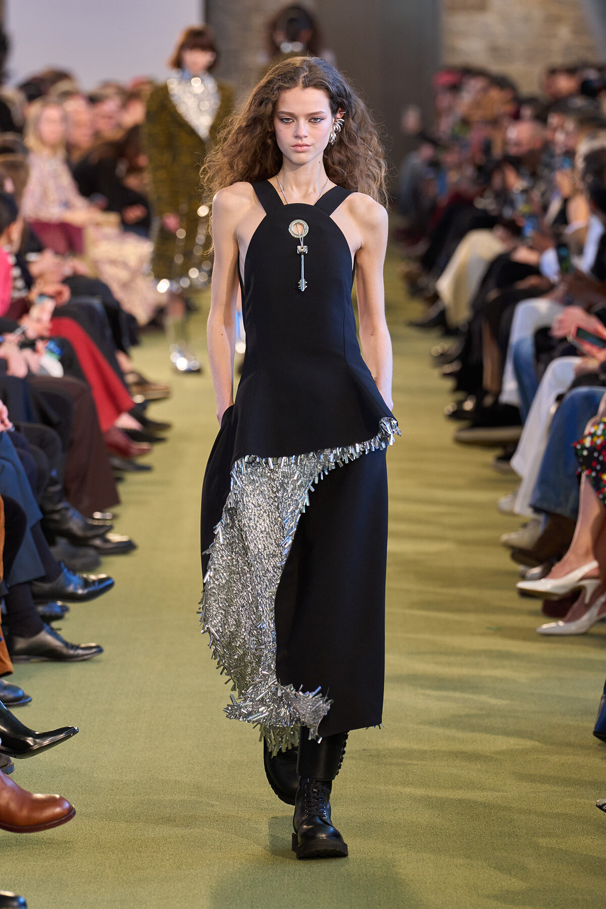 Model walking the runway in a black sleeveless dress with a silver fringe detail cascading down the front-left side of the skirt edge.