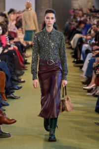 Model walks a fashion show wearing a glittery dark-green blouse tucked into a burgundy leather midi skirt with a wide belt, teal boots, and a fur handbag.