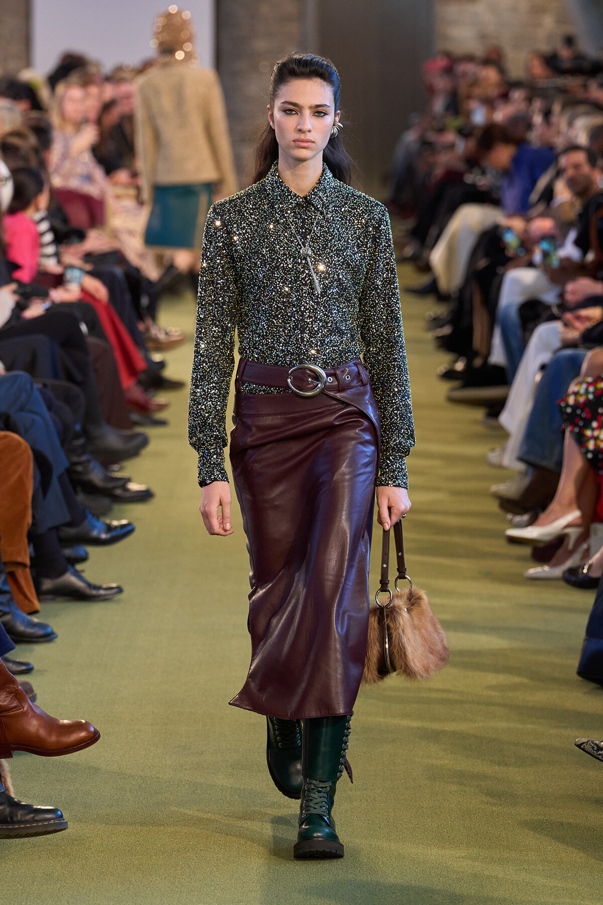 Model walks a fashion show wearing a glittery dark-green blouse tucked into a burgundy leather midi skirt with a wide belt, teal boots, and a fur handbag.